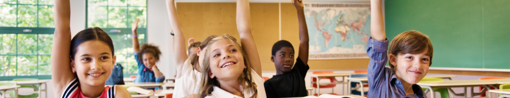 Diverse group of elementary school students sitting at desks, smiling and raising their hands high above their heads.