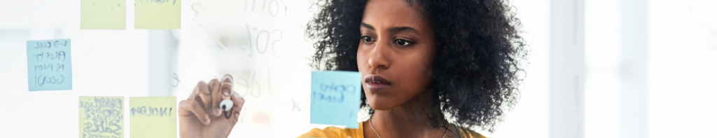 An African American woman brainstorms plans by writing with a dry-erase marker on a glass wall that also includes sticky notes.