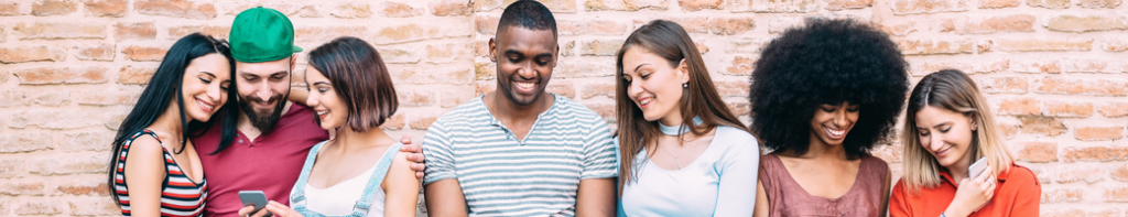 Diverse group of young adults stands against a brick wall, smiling and looking at their phones, taking selfies, and talking.