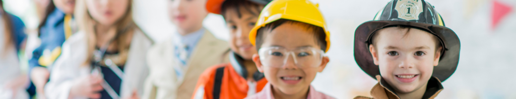 Diverse group of young children wearing career uniform costumes standing in a line. At the forefront is a boy dressed as a firefighter and a boy dressed as a construction worker.