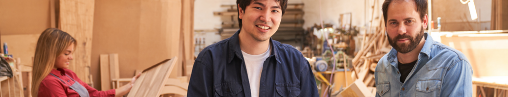 Asian-presenting male teen standing next to bearded white man in a workshop with a teen female working in the background.