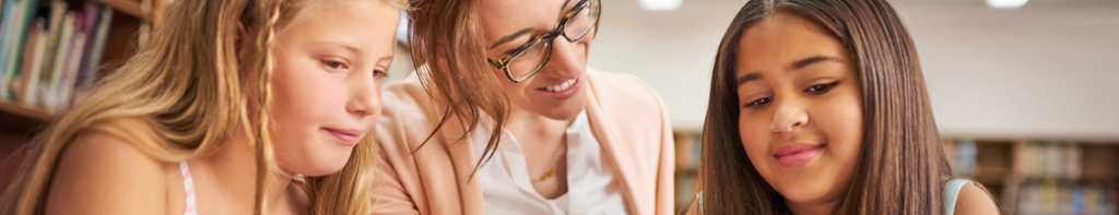 Female teacher smiles as she assists two diverse female students.