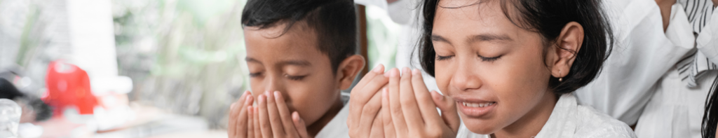 Young male and female Asian-presenting children with their eyes closed and hands raised in gratitude.