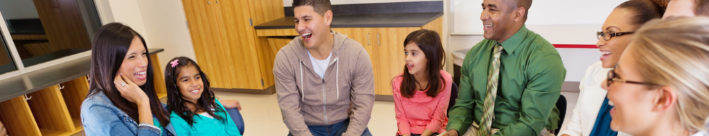 Diverse group of teachers, parents, and elementary students sit in a discussion circle smiling and laughing.