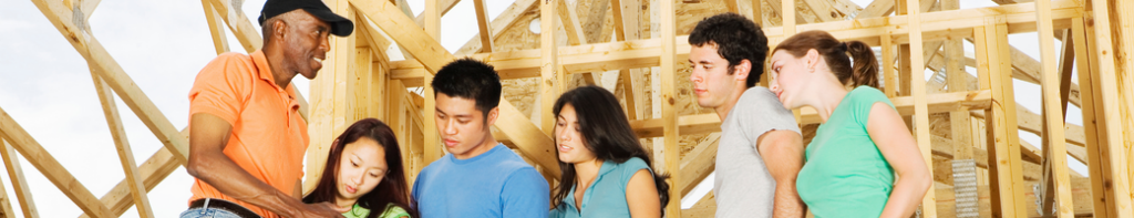 Diverse group of high school students listen to instructions being given to them by a Black male construction foreman. The frame of a building is in the background.