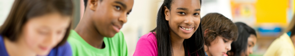 Diverse group of middle school students work side by side at a table. A Black female student smiles at the camera.