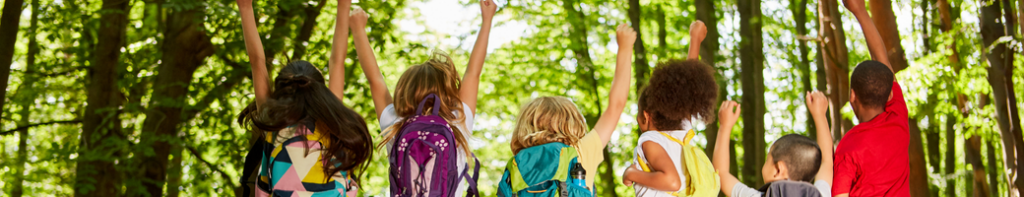 Diverse group of elementary students facing away from the camera jumping with their arms stretched up in victory and excitement on a field trip in a green wooded area.