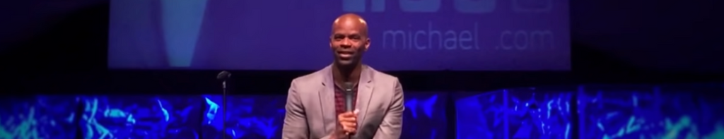 Comedian Michael Jr., a Black man, talking into a silver handheld microphone against a blue background and projector screen.