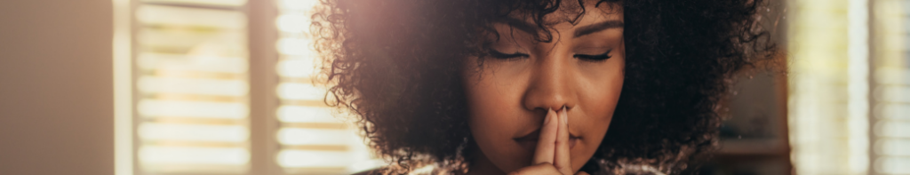 Black woman with natural hair and sunlight radiating through window blinds behind her has her eyes closed and index fingers pressed together against her closed mouth in a moment of calm mindfulness.