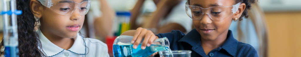 Two elementary students wear safety goggles as they compete a science activity using flasks filled with blue liquid.