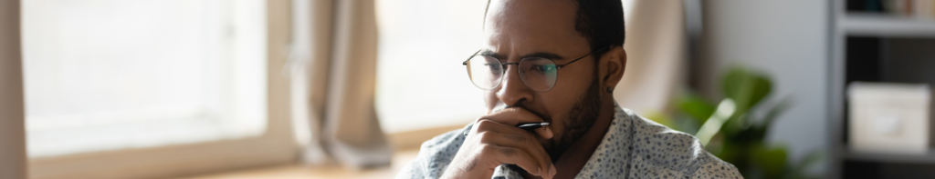 A man with dark hair and glasses rests his chin on his hand with a contemplative expression on his face.