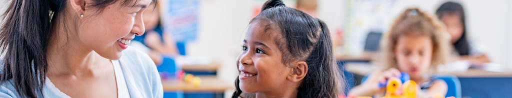 Asian female teacher smiles at a young dark-haired female student smiling up at the teacher.