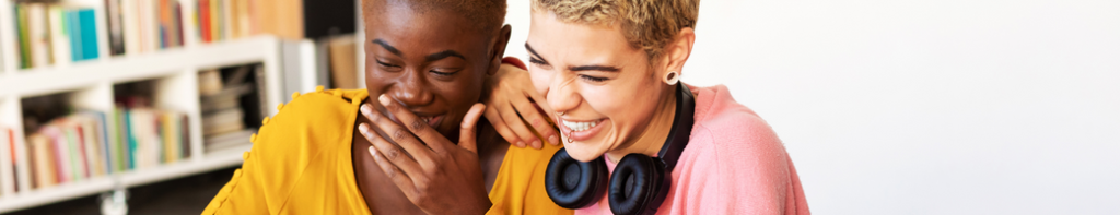 Two female high school students with short hair laugh together. One has a hand over her mouth indicating their laughter is mixed with embarrassment.