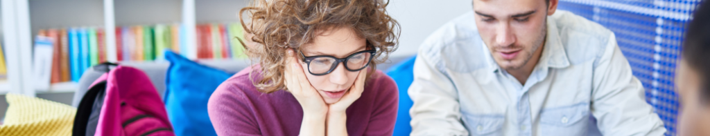 White female high school student with short curly hair and black frame glasses rests her chin in her hands with a puzzled expression on her face.