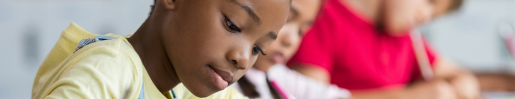 Black female student with her head down while handwriting. Other students blurred in the background are also handwriting.