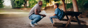 A male teacher squatting down to talk with a male student, whom looks upset, The student has a backpack on and is sitting on a bench of a picnic table.