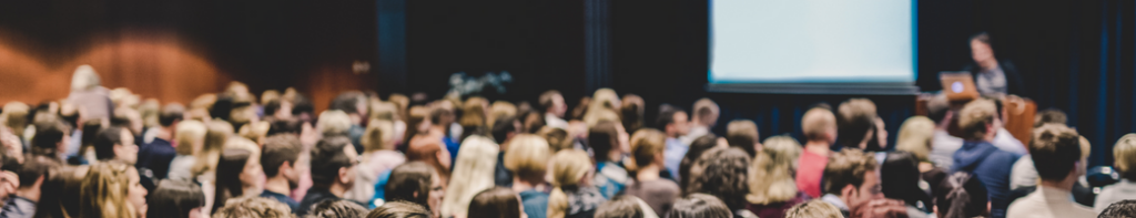 Diverse audience sitting in an auditorium with a large projection screen, watching a presenter.